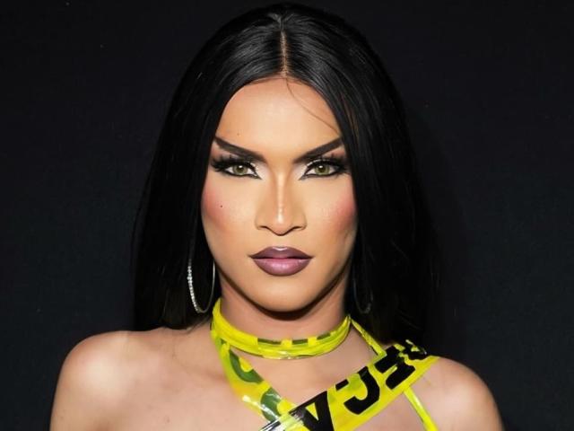 AgathaRodriguez has black hair, green eyes, dark makeup, and a yellow "caution" tape necklace. She's wearing purple lipstick and silver hoop earrings, with a confident expression.