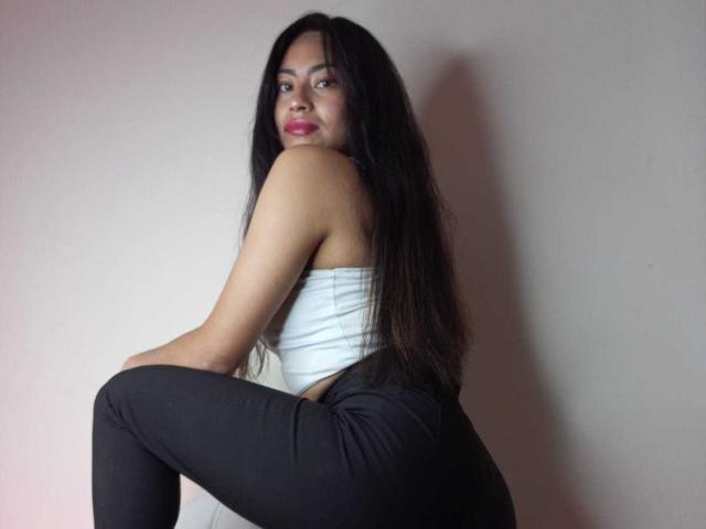 EmilyMitchell with long black hair, wearing a white tube top and black pants, sitting with one leg up, showing her ass, looking over her shoulder with a slight smile.