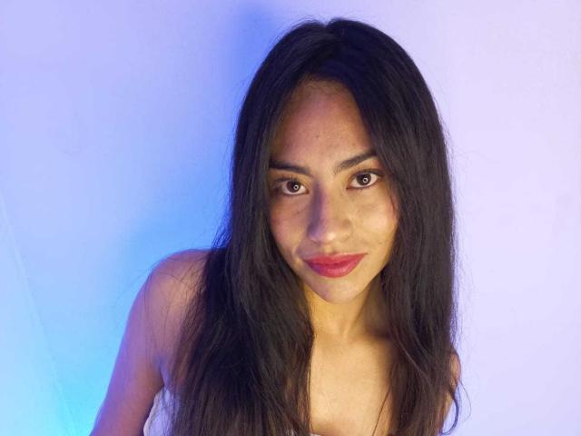 Emily Mitchell tem cabelos pretos longos, olhos castanhos e batom rosa, olhando levemente para baixo com um sorriso discreto, mostrando seus ombros nus. Ela tem um tom de pele marrom claro..