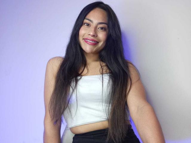 Emily Mitchell, com longos cabelos pretos, sorrindo, vestindo um top branco de alças finas, que revela seus pequenos seios, e calças pretas, mostrando uma pequena tatuagem em seu braço direito..