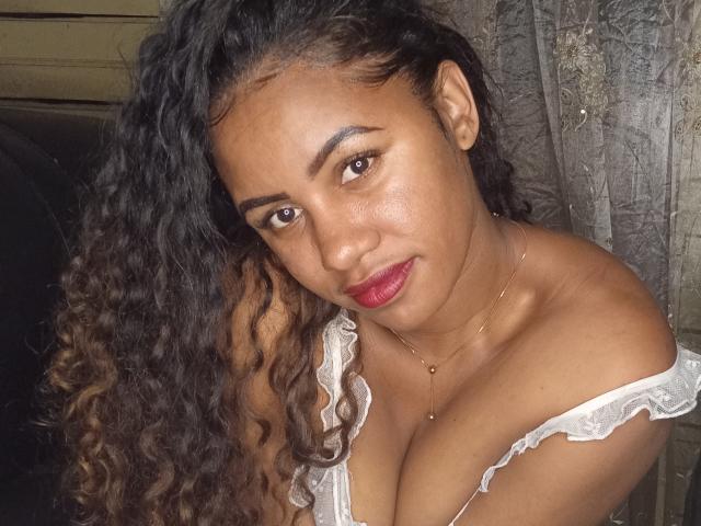 LayahCoquine with curly brown hair, medium brown skin, red lipstick, and white lace top, showing her tits, looking at the camera with a slight smile, wearing a delicate necklace.