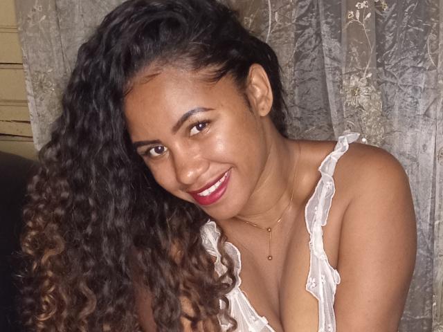 LayahCoquine has dark curly hair, brown skin, and is smiling with red lipstick, wearing a white ruffled top that shows her small tits. She's sitting with a necklace.