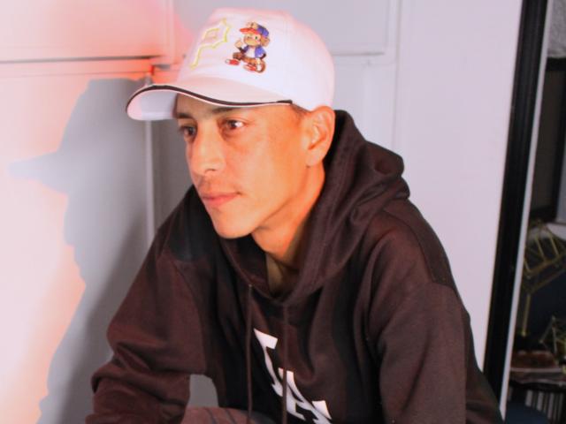 LatinDreams69 with a light skin tone, wearing a white cap with a cartoon, brown hoodie, and short brown hair, leaning against a white wall, looking slightly to the side.
