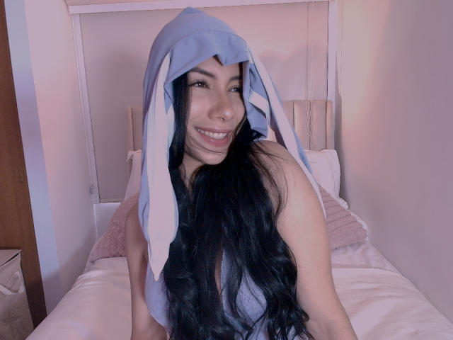 ArianaRogers with long black hair, wearing a blue bunny hood, smiling, showing her teeth, and sitting on a bed, her tits visible under the hood.