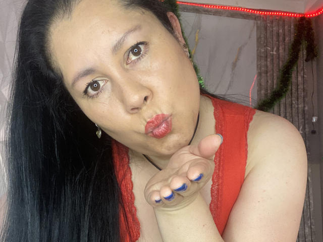 MarianaSmitt met lang zwart haar, een rode top van kant, volle rode lippen, blauwe nagellak, en een hand in de buurt van haar gezicht, die een kusbeweging maakt, waarbij haar borsten zichtbaar zijn..