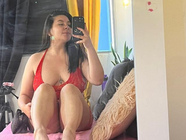 EmaJames with large tits in a red lace bra, sitting on a bed, taking a mirror selfie with a phone, knees up, black hair, yellow curtain in background.
