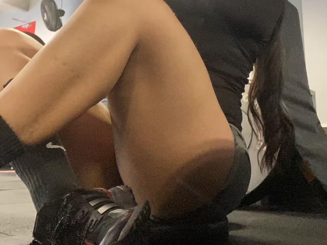 MiaDuboisX is squatting, showing her tan, muscular ass in black shorts and white socks, with long brown hair, black tank top, and black sneakers, in a dimly lit room.