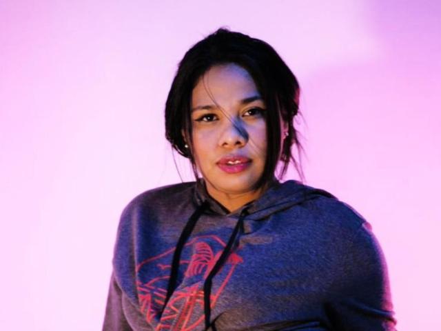 VanessaFory with black hair, medium brown skin, wearing a gray hoodie with red design, looking slightly to the side, pink and purple lighting. Boobs, ass not visible.