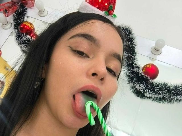 LissyCarter with black hair, wearing a Santa hat, has a green candy cane in her mouth, tongue out, and eyeliner, in a selfie with Christmas decorations in the background.