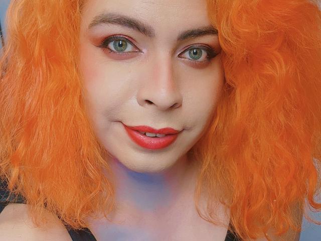 A TsFoxCandy tem cabelos encaracolados laranja vibrante, olhos verdes, batom vermelho e um leve sorriso, revelando um rosto adorável e sexy, com maquiagem discreta e um tom de pele claro..