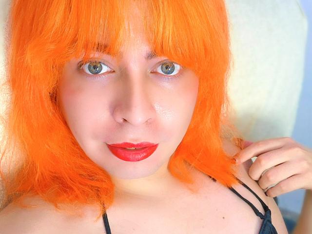 TsFoxCandy mit leuchtend orangefarbenem Haar, blauen Augen und rotem Lippenstift, trägt einen schwarzen Träger, berührt ihre Schulter und zeigt ein leichtes Lächeln. Sie hat ein glänzendes Gesicht..