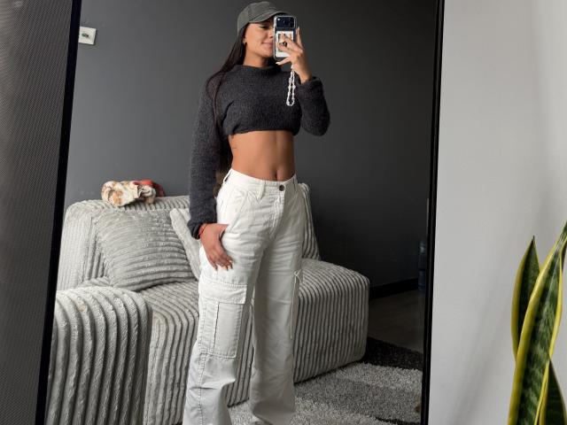 ThaliaAres, avec un ventre plat, portant un débardeur gris, un pantalon cargo blanc et une casquette, prend un selfie devant un miroir, montrant ses fesses et ses abdominaux tonifiés..