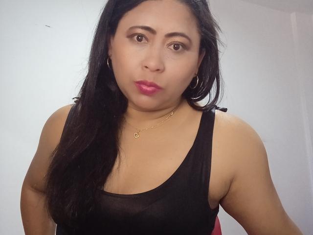 MiaSalomeChaude has tan skin, black hair, pink lipstick, hoop earrings, a black tank top, and a gold necklace. Her boobs are small to medium. She's making a pouty face.