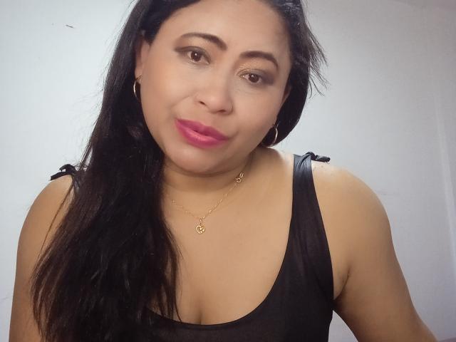 MiaSalomeChaude has brown eyes, black hair, pink lipstick, and a black tank top showing her tits. She's wearing a gold necklace and hoop earrings.
