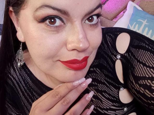Gaiax69 with red lipstick, black mesh top with cut-out fingers, long nails, silver earrings, and a slight smile. Brown eyes, light skin, nose ring, and black hair.