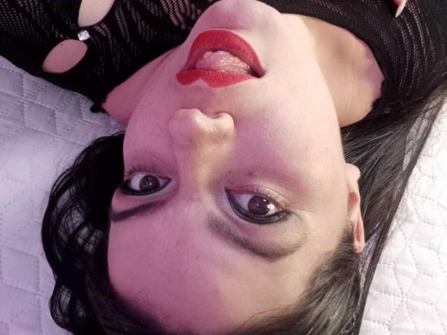Gaiax69's face, upside down, with red lips, tongue out, black mesh top, dark eyebrows, brown eyes, and black hair on a white quilted bed.