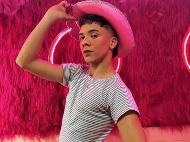 BlakeNoir with curly brown hair, wearing a striped shirt, pink cowboy hat, holding hat with left hand, pink neon lights in background, slight smile, necklace, looking to the side.