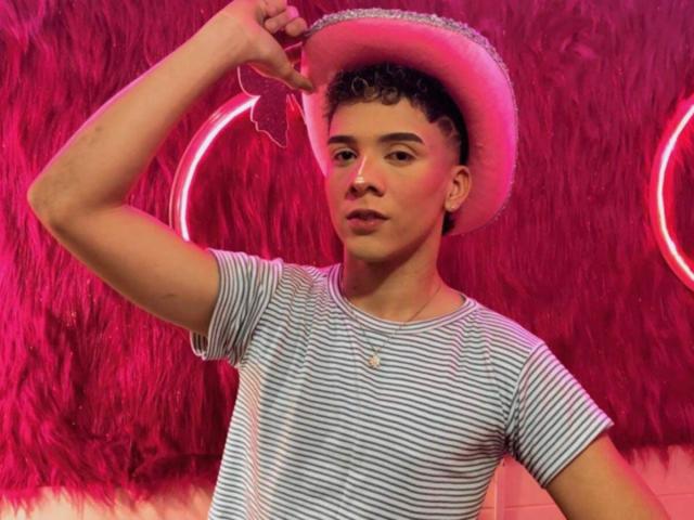 BlakeNoir wears a pink cowboy hat, striped shirt, and a necklace, raising his arm, with curly brown hair, a cute face, and a confident look.