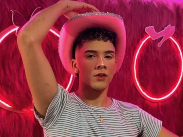 BlakeNoir with curly brown hair, wearing a white felt hat, striped shirt, and earring, holding the hat with his right hand, neon pink lighting, and a relaxed expression.
