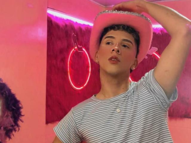 BlakeNoir wearing a pink hat, striped shirt, with one arm raised, looking slightly to the side, pink neon ring in background, short curly brown hair, soft pink lighting.