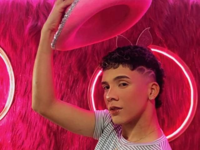 BlakeNoir, holding a pink, glittery hat, with short curly hair and a striped shirt, looking up, neon lights in the background. Handsome, light skin, cute, semi-profile.