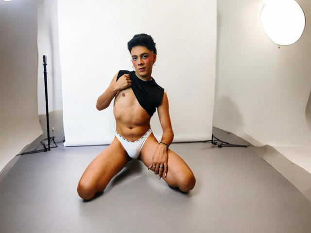 BlakeNoir, kneeling, with short black hair, lifting a black top to expose small tits, wearing white panties, on gray floor, under studio lights, looking at camera.