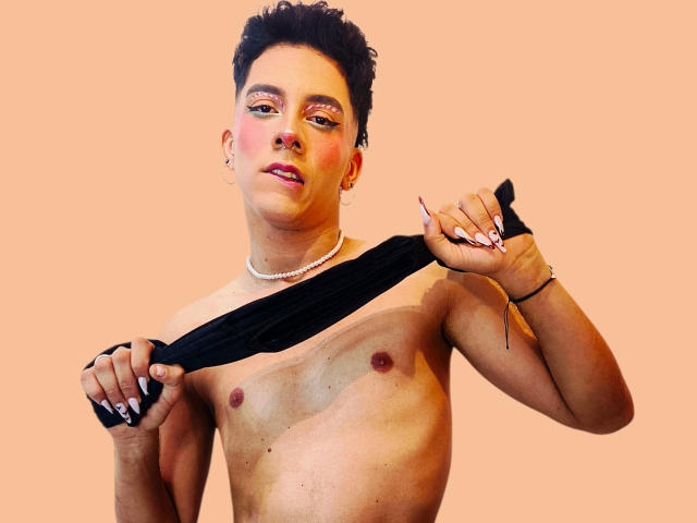 BlakeNoir, shirtless, holding black shirt, pale peach background, short dark curly hair, nose ring, pearl necklace, pinkish blush, small nipples, ring on left hand, pink nails.