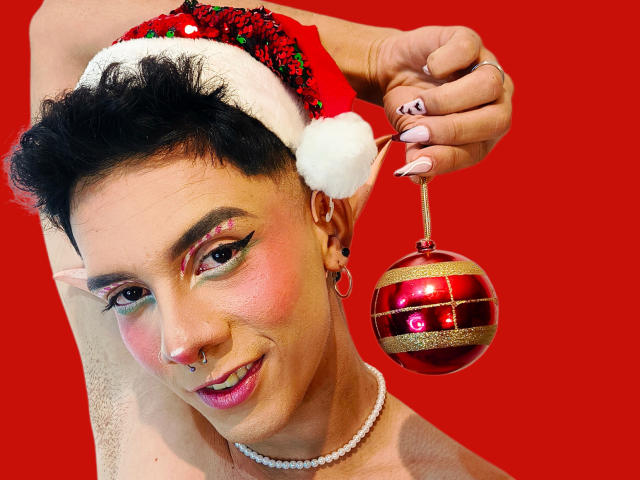 BlakeNoir with short black hair, wearing a red Santa hat, holding a red and gold Christmas ornament, has pink nail polish, a nose ring, and a white pearl necklace.
