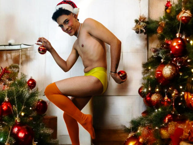 BlakeNoir, shirtless, wearing yellow underwear and orange thigh-high socks, holds Christmas ornaments, wearing a Santa hat, standing between two decorated Christmas trees, smiling.