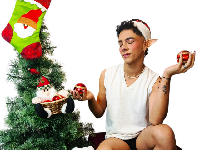 BlakeNoir, with elf ears, white tank top, and red headband, holds Christmas ornaments, sitting by a decorated tree with a green sock and a gnome. White background.