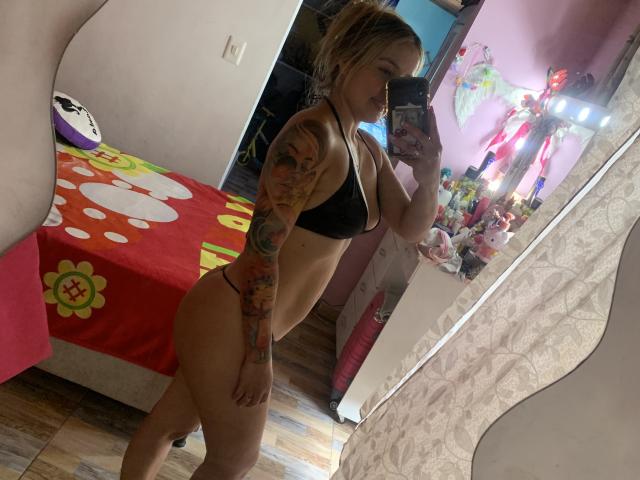 SaritaCaramelo with tattoos, black bra, and thong, standing in profile, taking a mirror selfie, showing her ass and small tits, in a pink-walled bedroom.