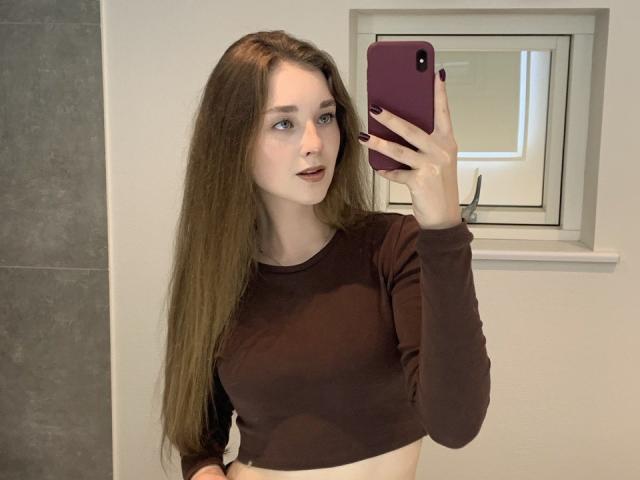 AlisaBlisss with long brown hair, wearing a brown long-sleeve crop top, holding a purple phone, taking a mirror selfie, showing her tits, and smiling slightly.