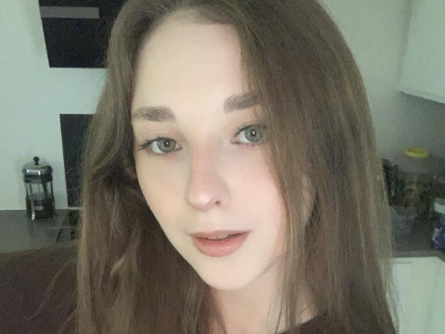 AlisaBlisss has pale skin, green eyes, and long brown hair. She's looking at the camera with a slight smile, showing her pink lips. She's wearing a black top.