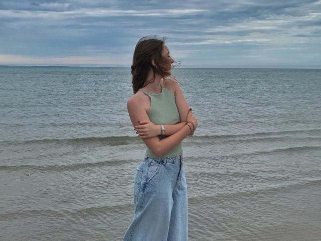 AlisaBlisss standing by the ocean, arms crossed, wearing a green tank top and high-waisted blue jeans, with her brown hair blowing in the wind.