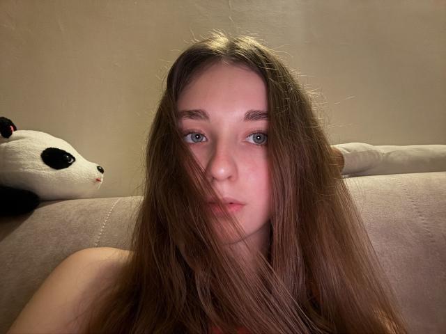 AlisaBlisss has long brown hair, blue eyes, and a slightly flushed face, sitting on a beige couch with a panda pillow in the background. Her tits are not visible.