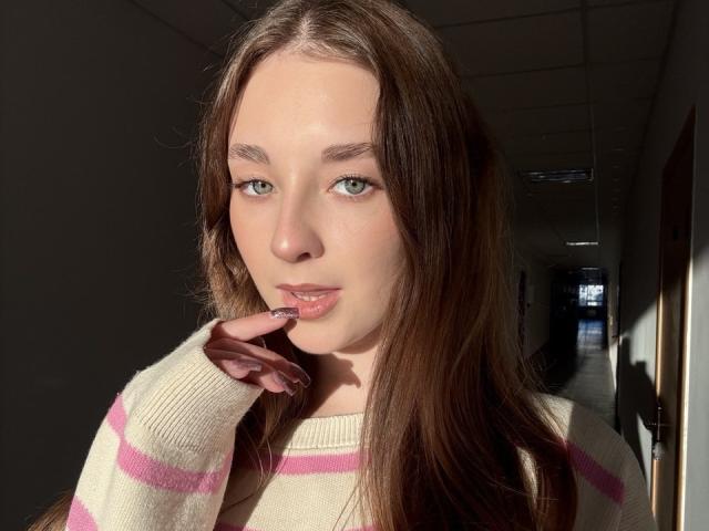 AlisaBlisss with long brown hair, green eyes, and pink-striped sweater, lightly biting her finger, sunlight on her face, shadowed hallway in the background.