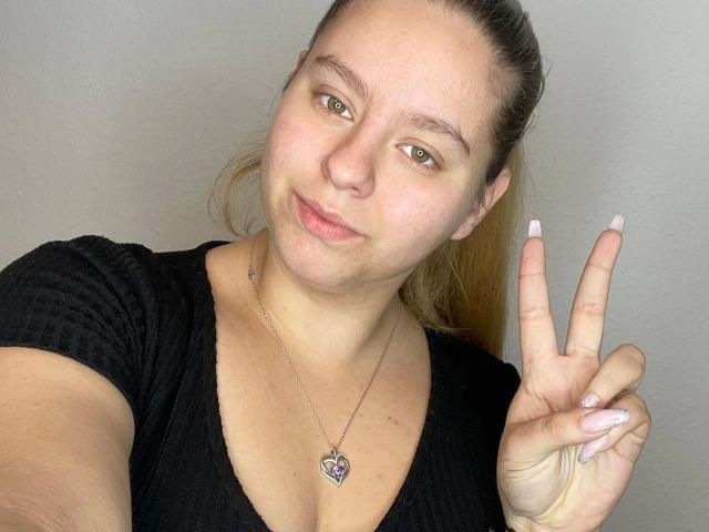 MiaLou05-hot has light skin, green eyes, and a slight smile. She's wearing a black ribbed shirt, a heart necklace, and making a peace sign with her right hand.