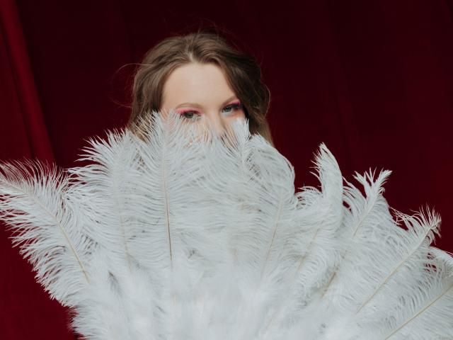 MariaRobinson's face is partially hidden by a bunch of white feathers, with just her pink-eyed, brown-haired face visible, set against a red background.