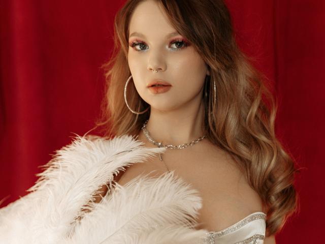 MariaRobinson has wavy, auburn hair and wears large, silver hoop earrings. Her makeup includes pink eyeshadow and red lipstick. She holds a white feathered garment that partially covers her bare shoulders. A silver chain necklace rests on her neck. The red background contrasts with her pale skin and the white feathers. Her blue eyes are focused and slightly parted. The image is detailed and has a soft, realistic texture.