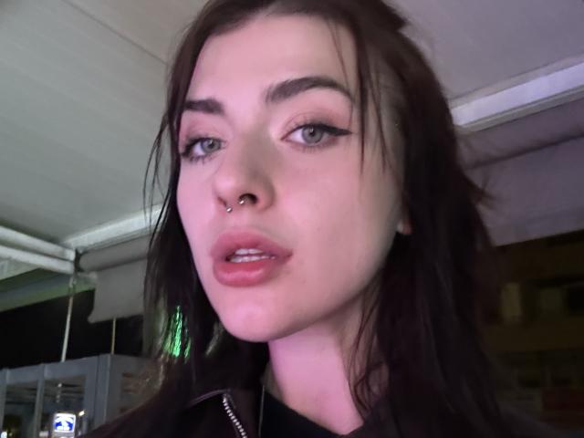 DarinaIngrid has pale skin, dark brown hair, a nose ring, and full pink lips. She's looking at the camera with half-closed eyes, wearing black makeup.