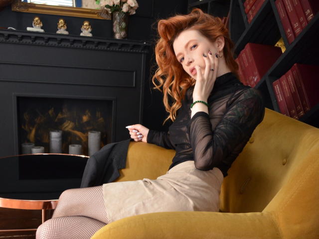 MeganSolar with red hair, black sheer top, beige skirt, fishnet stockings, yellow chair, hand on cheek, looking at camera, black jacket on arm, bookshelf, fireplace, candles.
