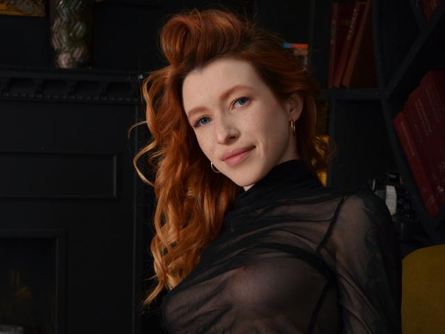 MeganSolar, avec ses cheveux roux, ses yeux bleus et ses taches de rousseur, porte un haut noir transparent qui révèle ses seins. Elle sourit, regardant l'appareil photo, avec des cheveux ondulés..