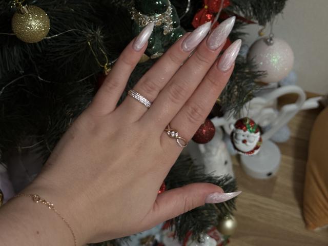 La mano di SugarrX, con unghie lunghe, rosa e scintillanti, che indossa due anelli e un delicato braccialetto, tocca un albero di Natale decorato con ornamenti rossi, dorati e bianchi..