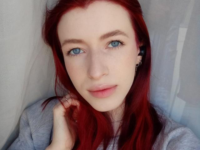 MeganSolar with red hair, blue eyes, and pale skin, wearing a gray top, looking at the camera with a slight smile, her hand near her face.