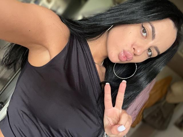 VanessaEsther with black hair, pink ombre tips, making a pouty face, wearing a black sleeveless top, large hoop earrings, flashing a peace sign, showing her tits.