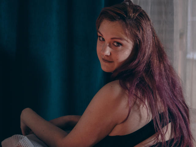 StellaSunfield with long, red-brown hair, looking over her shoulder, wearing a black top, showing her bare back and side-boob, sitting in a dimly lit room.