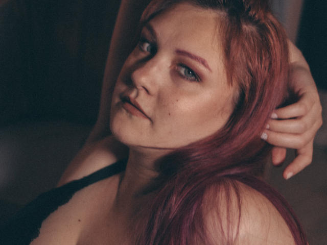 StellaSunfield with red hair, slightly parted lips, and a relaxed expression, her hand in her hair, wearing a black lace top, showcasing her bare shoulders and subtle freckles.