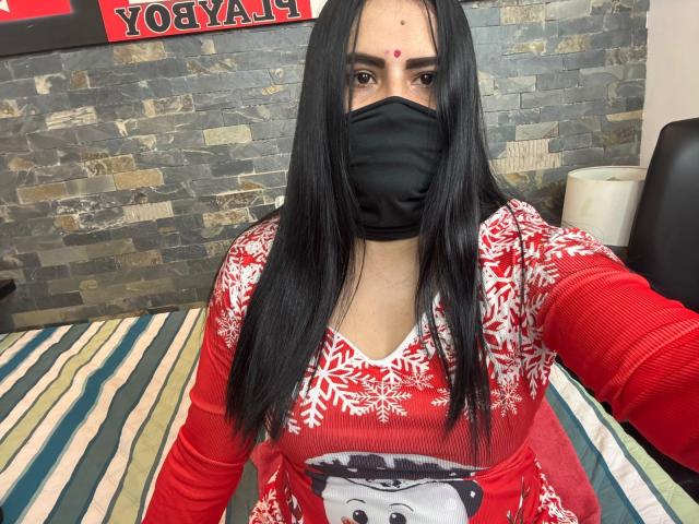 DaliaDubey with long black hair, red face mask, red Christmas sweater, white snowflake pattern, and a photo of a black and white face on the chest. She's wearing a red dot.