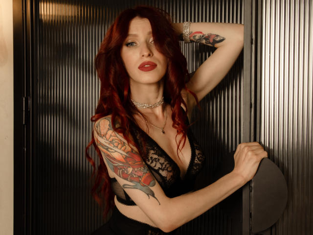 MeganSolar, avec ses cheveux roux, son haut noir en dentelle et ses tatouages, tenant une porte, portant un collier en argent, bras levé, montrant ses seins et une expression sexy et confiante..