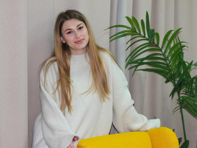 AlinaNathan with long blonde hair, white sweater, smiling, sitting on yellow chair, holding armrest, green plant to right, soft light, relaxed, cute, attractive, feminine, casual.
